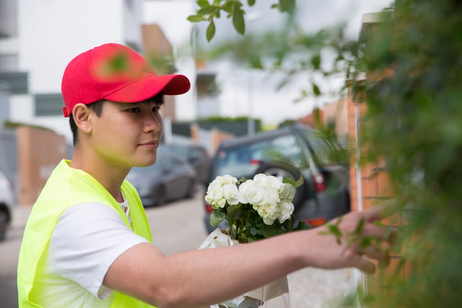 A young male florist wearing a red cap and a bright yellow high-visibility vest is delivering a floral gift. He is holding a bouquet of fresh white hydrangeas wrapped in white paper, with vibrant gree