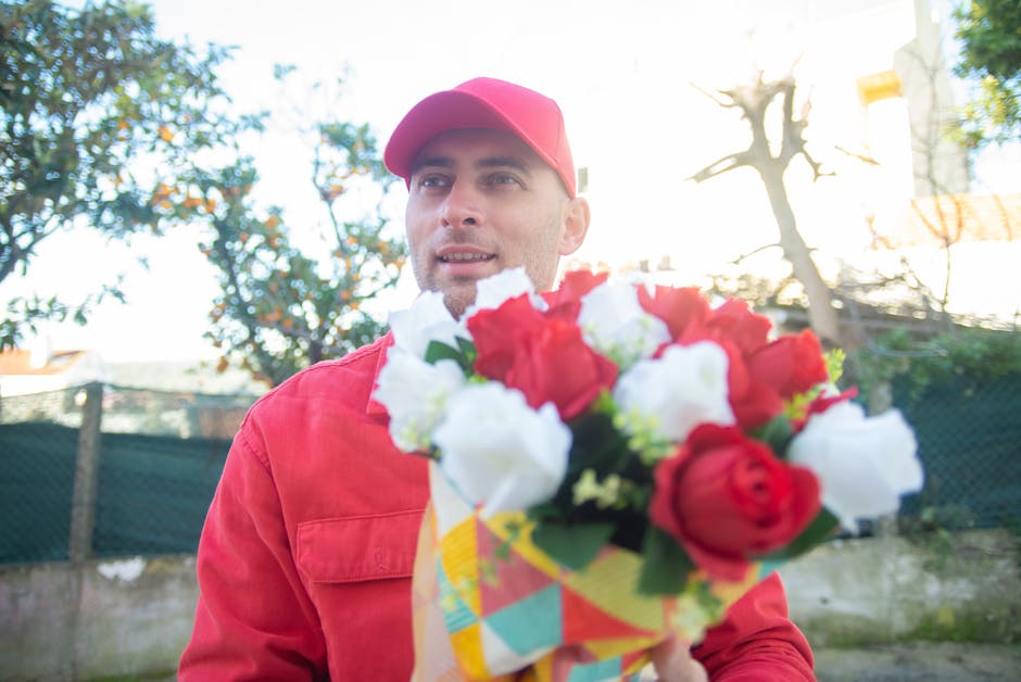 A male florist dressed in a red uniform and cap holds a vibrant floral bouquet in front of him outdoors, with a background of trees, a fence, and a partly cloudy sky illuminated by sunlight. The bouqu