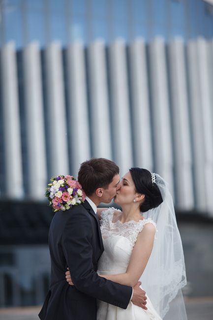 A bride and groom sharing a kiss outdoors in front of a modern building with vertical glass panels, the bride wearing a white lace wedding dress with lace cap sleeves and a veil, and the groom in a da
