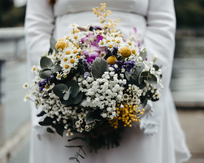 A person in a white outfit holding a large, vibrant bouquet of fresh flowers outdoors. The bouquet features a mix of white daisies, small white and yellow blossoms, purple orchids, dark purple statice