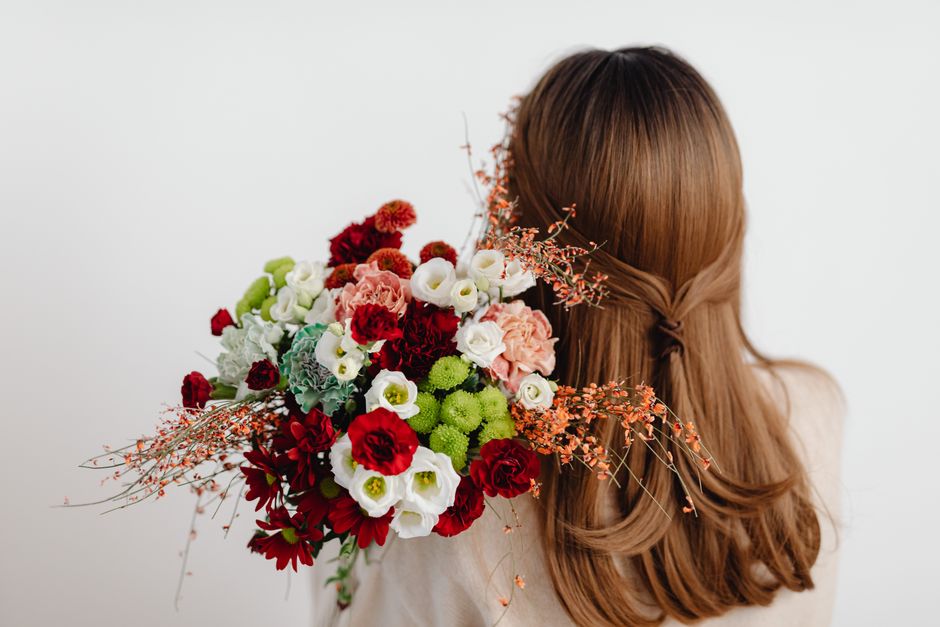 A woman with long, wavy auburn hair, partially styled with a twisted strand, holds a vibrant floral bouquet close to her shoulder. The bouquet features an assortment of fresh flowers, including deep r