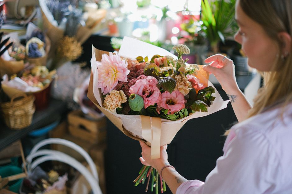 A woman with blonde hair, wearing a light pink shirt, stands in a floral shop holding a large, freshly arranged bouquet of mixed flowers. The bouquet features prominent pink roses, a large pale pink d